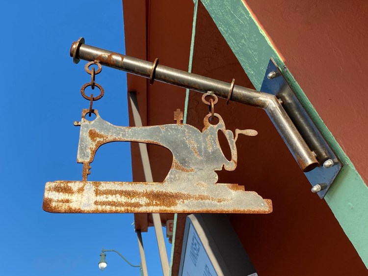 A rusted metal sewing machine cutout hangs from an angled bar on the outside wall of an alterations business.