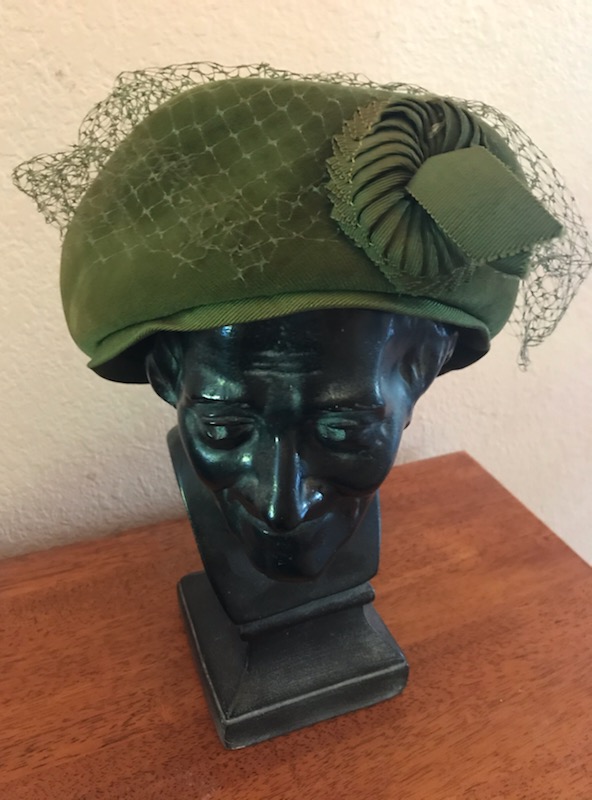 A green cloche hat with tousled green netting and a green grosgrain ribbon rosette sits on a black marble sculpted head.